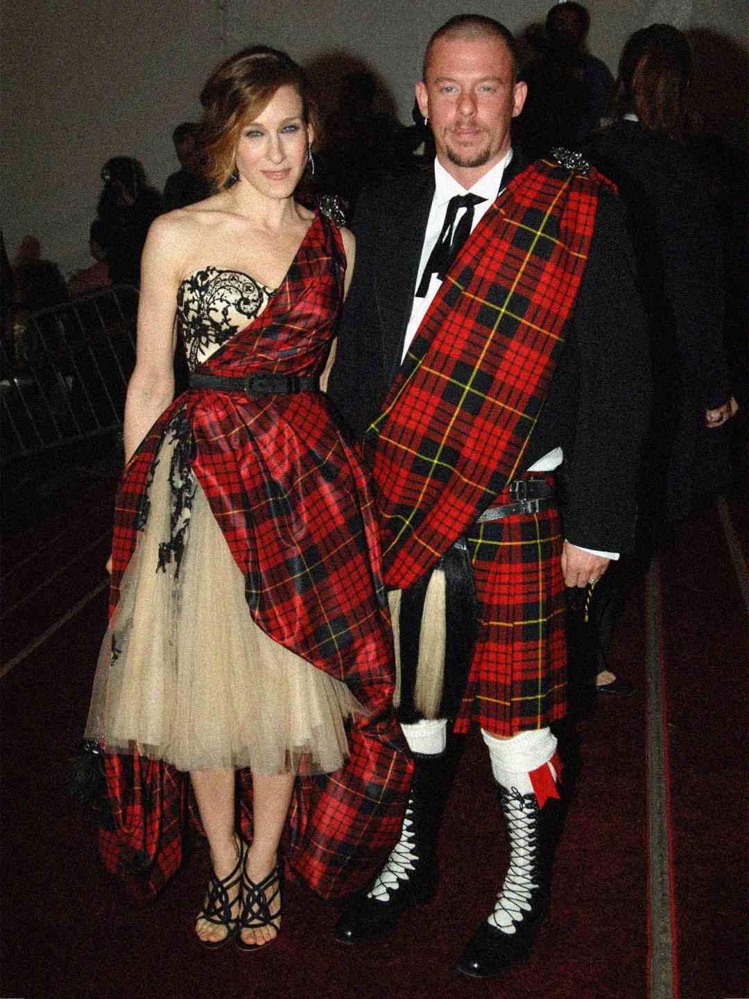 A woman in a strapless beige dress with a red tartan sash stands next to a man wearing a traditional Scottish kilt outfit with a matching red tartan pattern. Both are posing and smiling at an event.