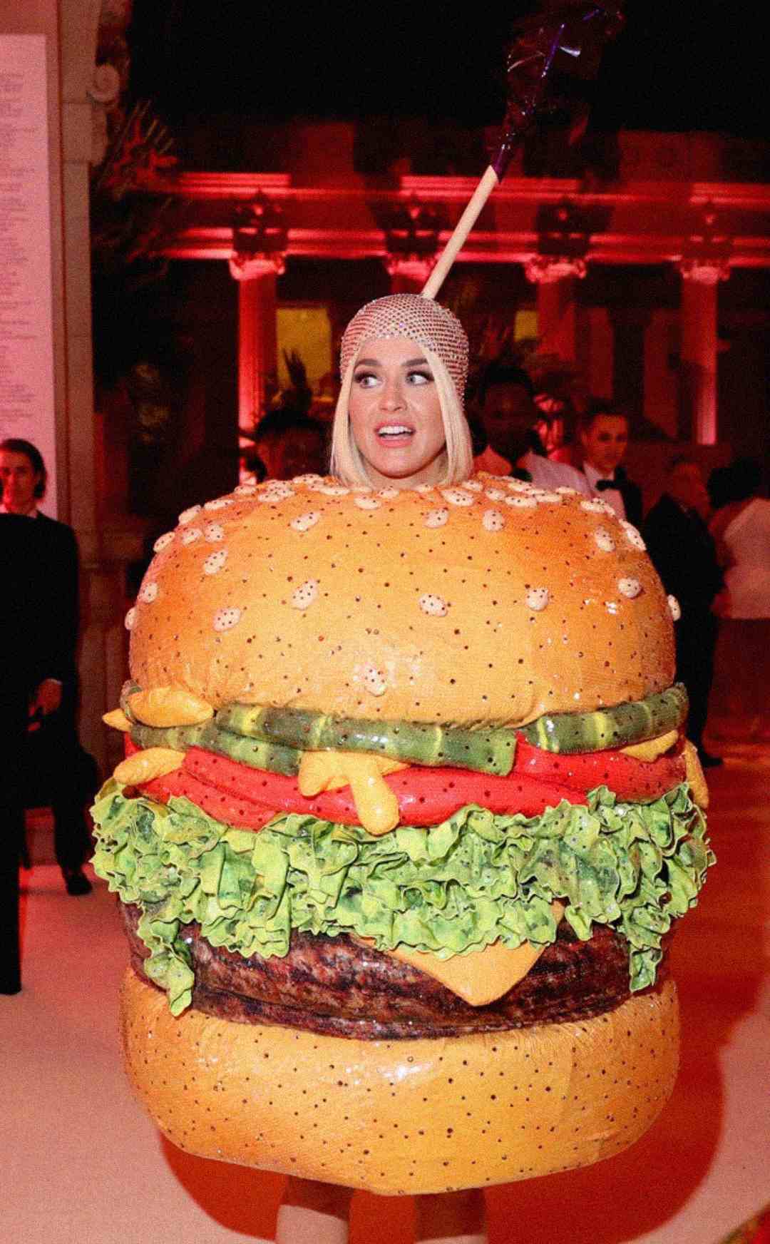 A woman wearing a large hamburger costume, complete with lettuce, tomato, cheese, and a bedazzled bun, stands indoors at a lively event with dramatic lighting and people in the background.