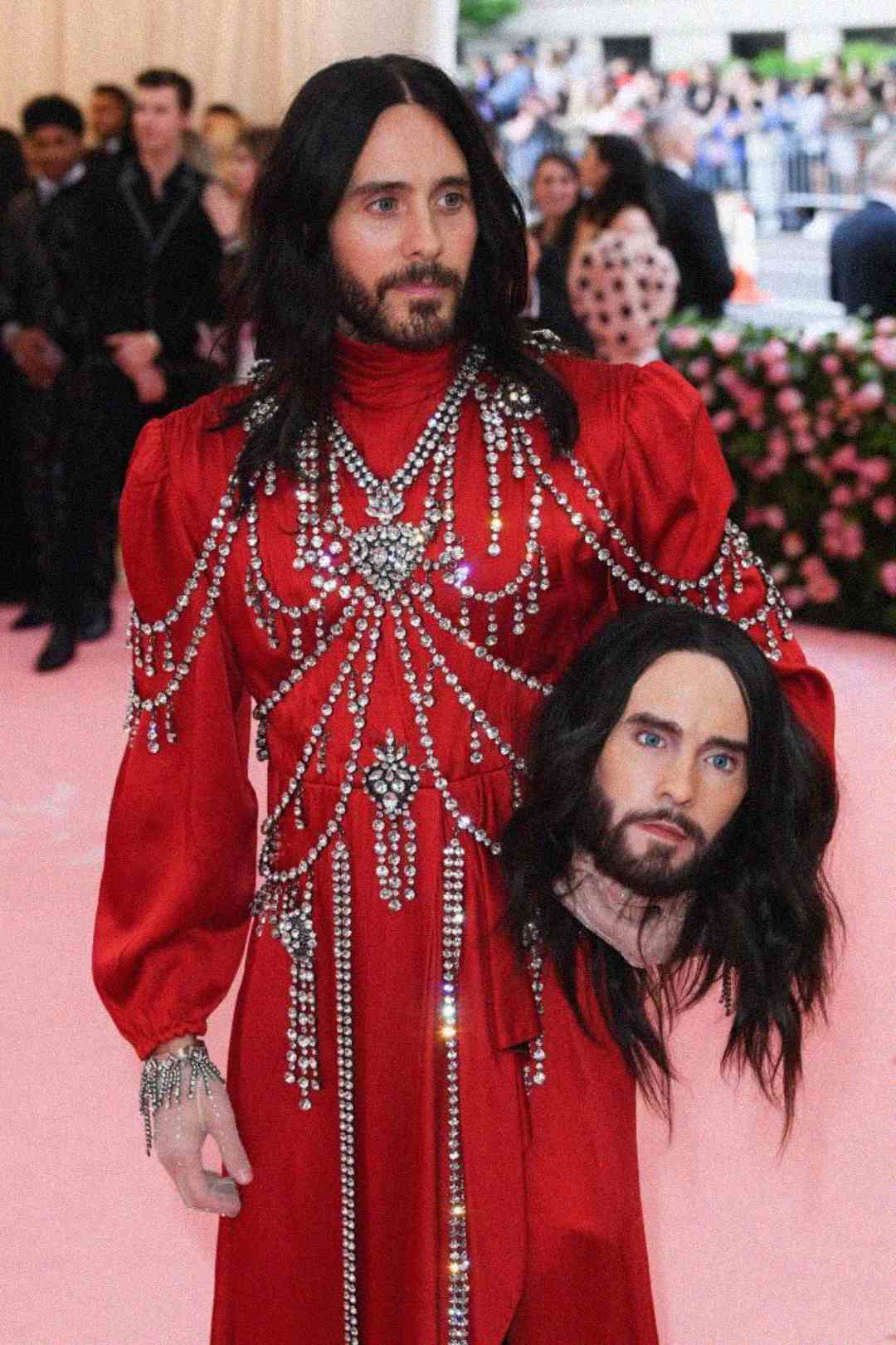 A person in a long red gown adorned with crystal chains stands on a pink carpet, holding a realistic replica of their own head. People in formal wear are blurred in the background.