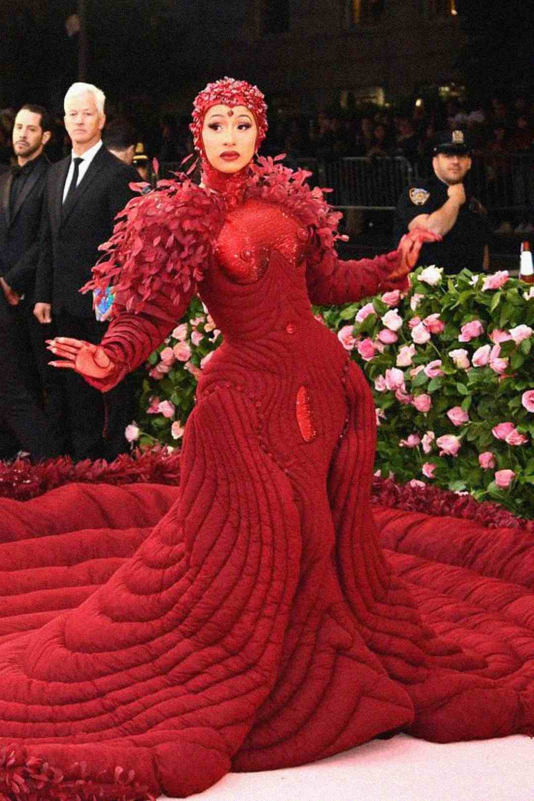 A woman poses on a red carpet in an extravagant, textured deep red gown with feathered shoulders, a dramatic train, and a matching headpiece. Pink flowers and people are visible in the background.