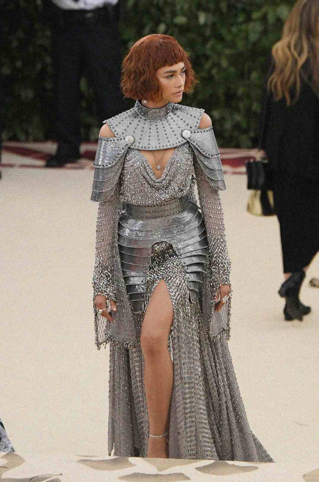 A woman wearing a silver, armor-inspired gown with intricate metallic detailing, shoulder plates, and chainmail sleeves. She has short, wavy auburn hair and is standing on a cream-colored carpet.