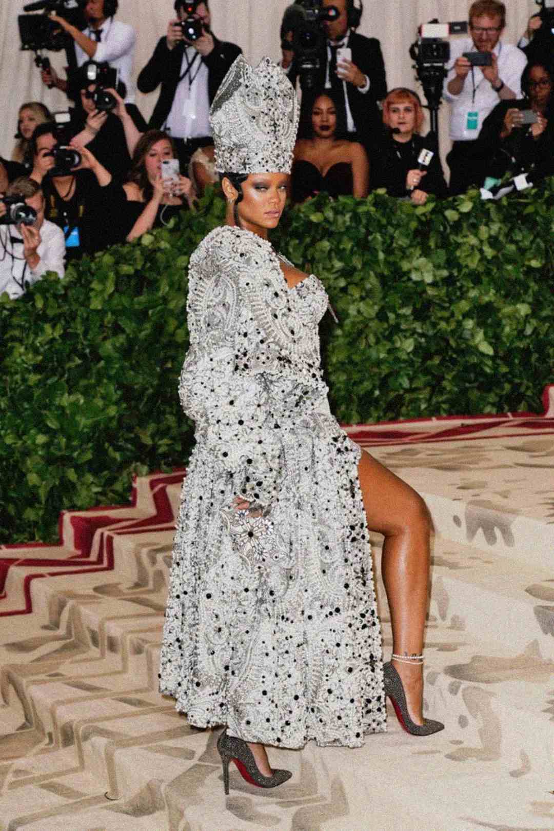 A woman poses confidently on a grand staircase, wearing an ornate, silver bejeweled gown and matching tall headpiece, surrounded by photographers and onlookers at a formal event.