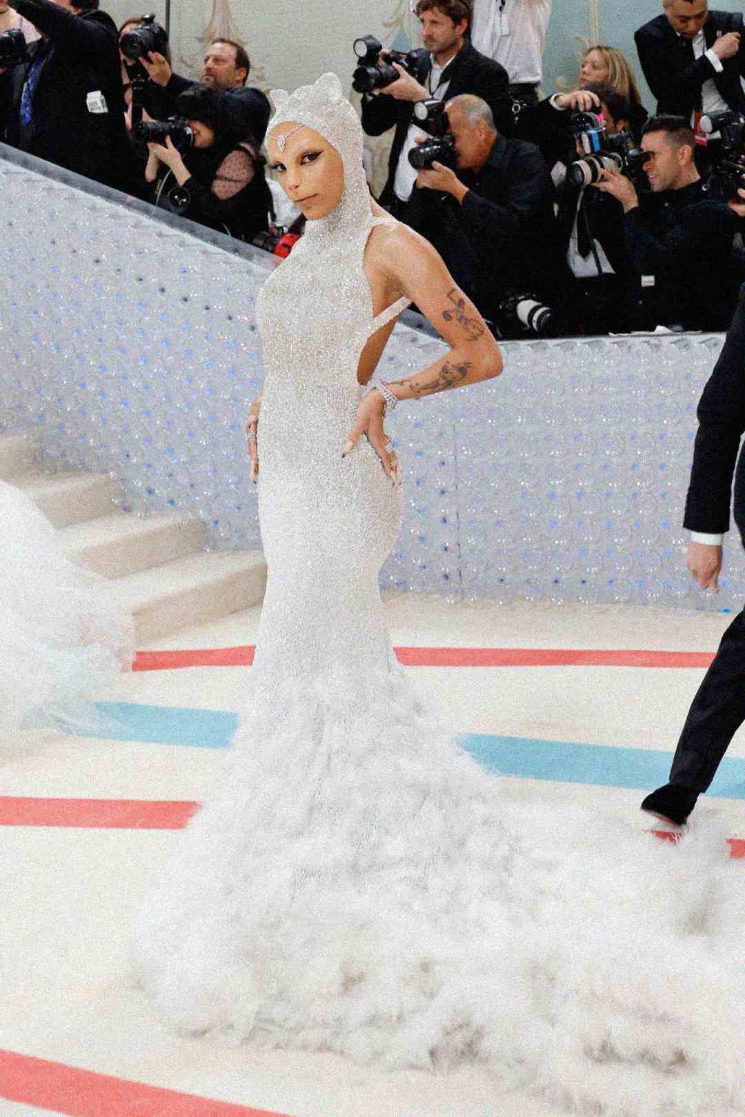 A person poses confidently on a carpeted stairway, wearing a sparkling, form-fitting white gown with a dramatic feathery train and an embellished headpiece, surrounded by photographers taking pictures.