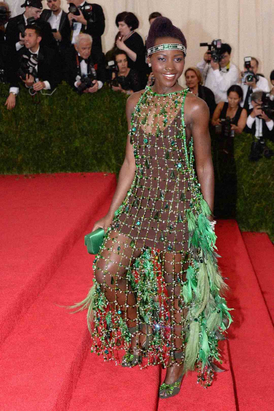 A woman poses on a red carpet wearing a sheer, beaded green dress adorned with feathers. She holds a green clutch and wears matching heels, with photographers and guests in formalwear in the background.