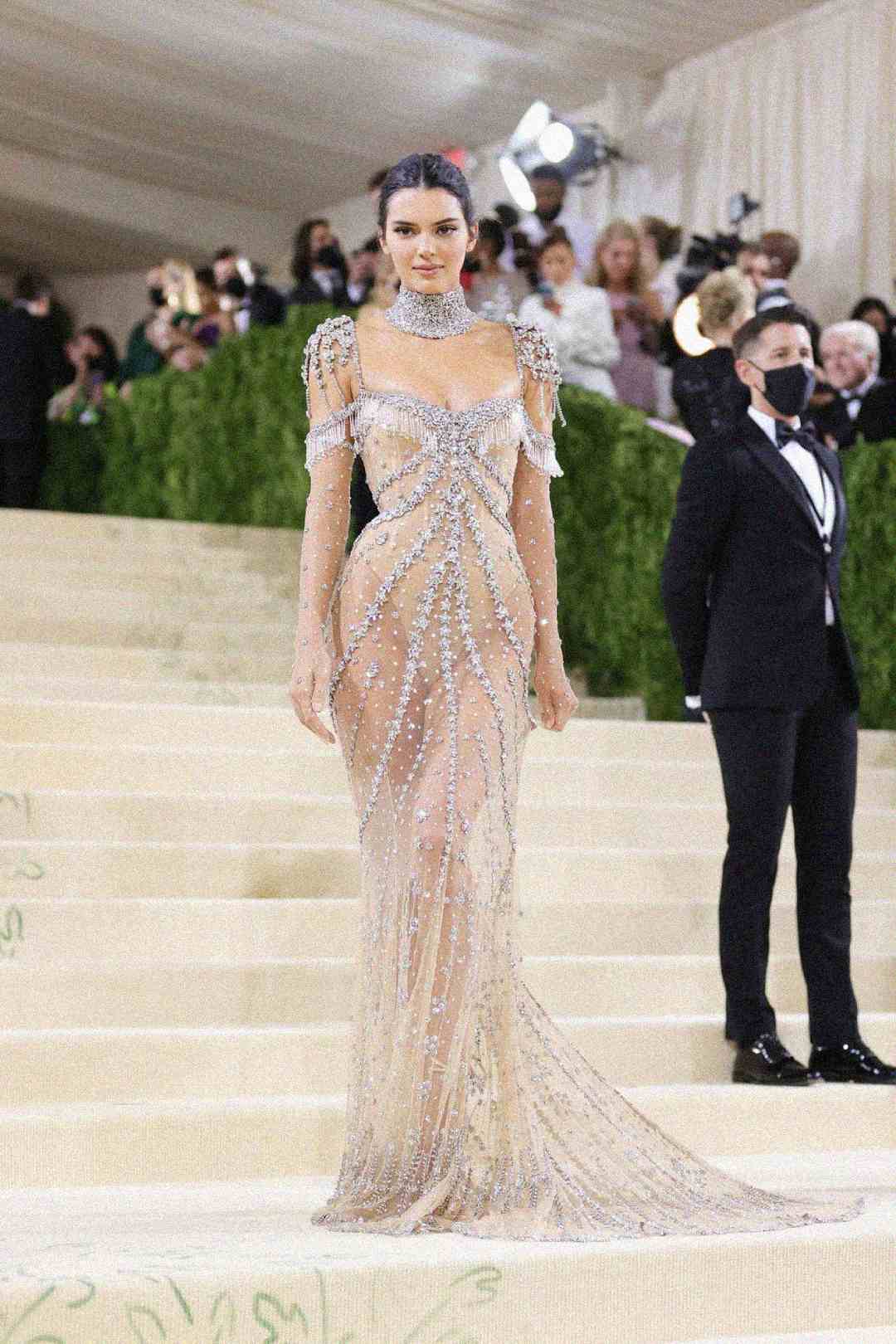A woman in a sheer, beaded, floor-length gown poses on a grand staircase at a formal event. She has her hair pulled back and is surrounded by people in elegant attire. A man in a tuxedo and mask stands nearby.