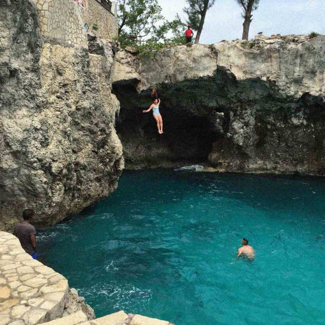 A person jumps off a rocky cliff into clear blue water, while another person swims below and a few others stand on the rocks watching. The scene is outdoors near a stone wall and natural cave.