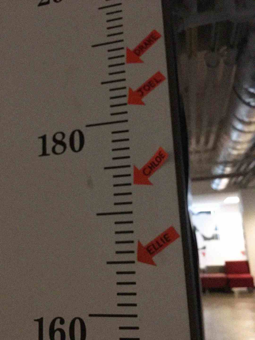 A height measurement chart shows marks near 180 cm labeled with red name tags: "DANIEL," "YOEL," "CHLOE," and "ELLIE." An industrial hallway with red chairs is visible in the background.