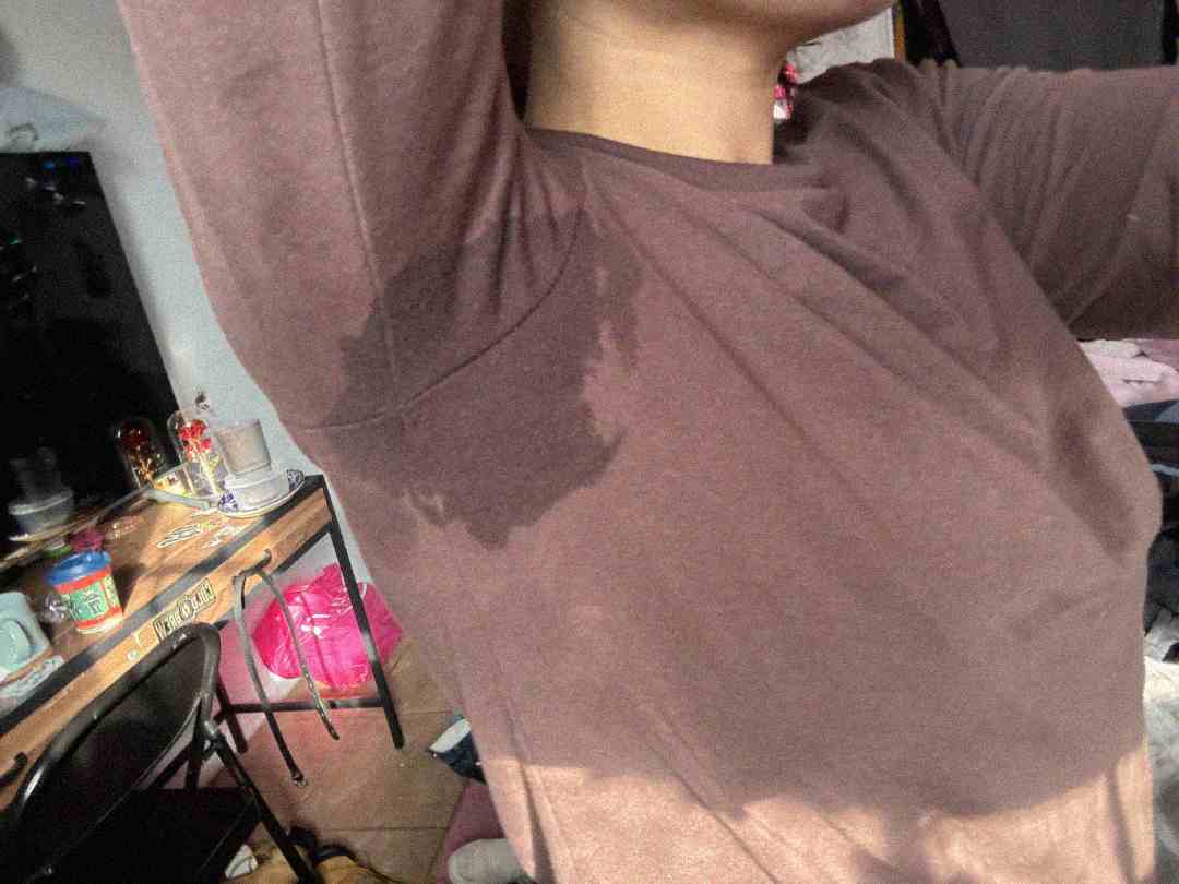 A person wearing a brown shirt with a large sweat stain under their arm raises their arm. The background shows a cluttered room with a desk, chair, and various items.