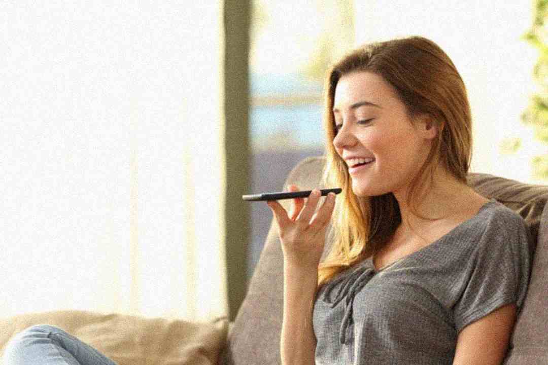 A young woman sits on a couch, smiling and holding a smartphone in front of her mouth, appearing to use a voice command or voice message feature. Soft light shines through windows in the background.