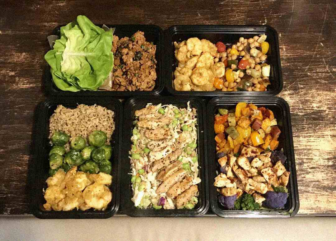 Six black meal prep containers filled with various healthy foods, including grilled chicken, rice, brussels sprouts, mixed vegetables, lettuce leaves, ground meat, and chickpeas, arranged on a rustic wooden table.