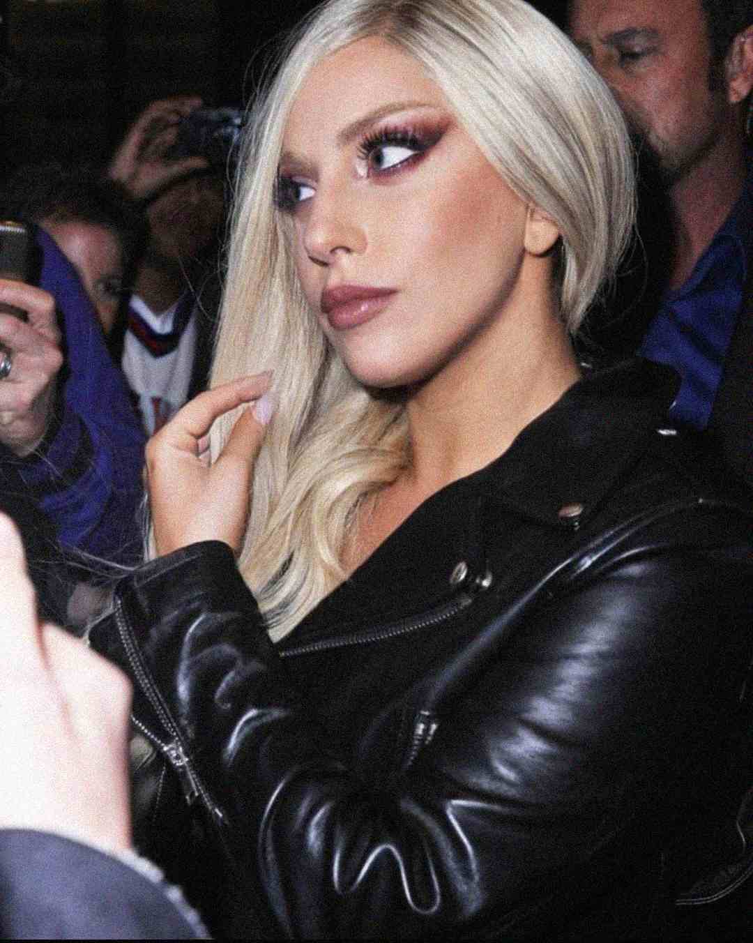 A person with long platinum blonde hair, dramatic makeup, and a black leather jacket looks to the side while surrounded by people and cameras in a crowded setting.