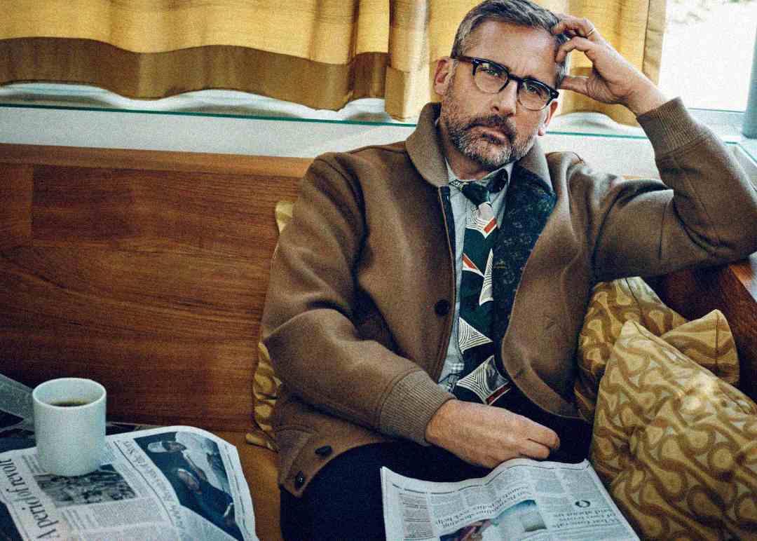 A man with glasses and a beard sits on a couch, resting his head on his hand. He wears a brown jacket and patterned shirt, with newspapers and a mug on the table nearby. Yellow curtains hang in the background.