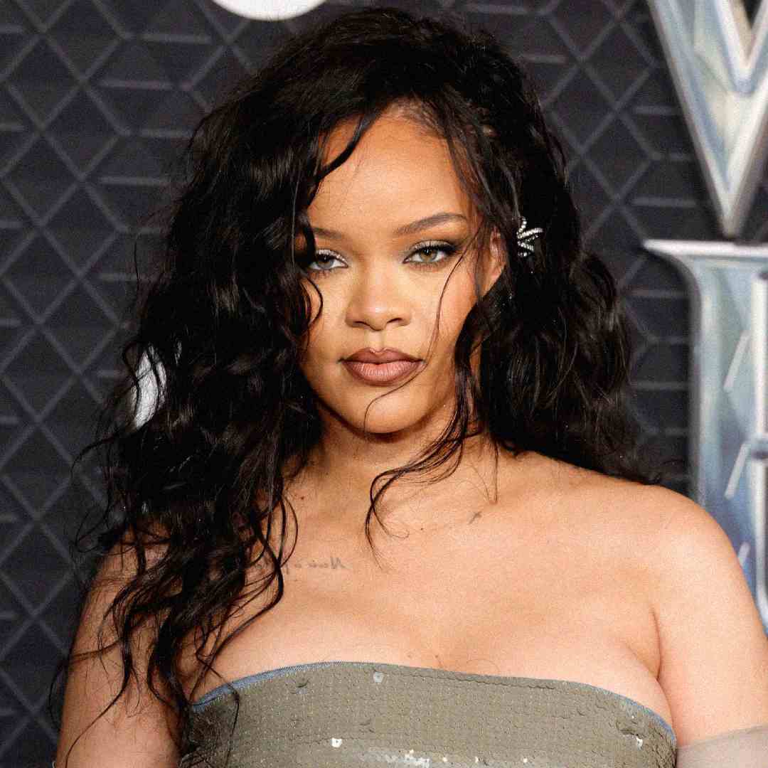 A woman with long, wavy black hair and green eyeshadow poses in front of a geometric, dark background. She wears a strapless, metallic dress and has a confident expression.