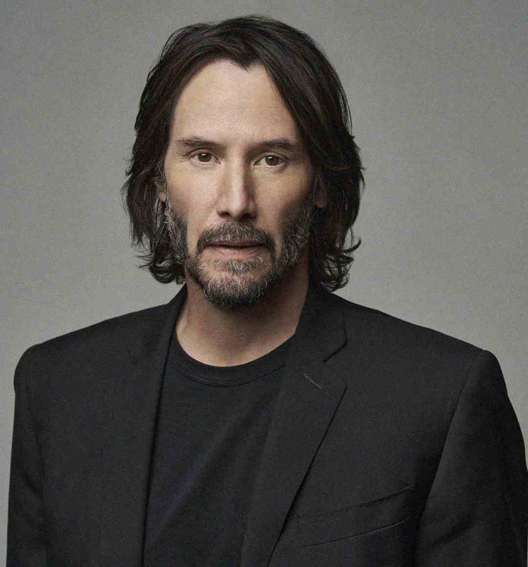 A man with shoulder-length brown hair and a beard wearing a black blazer over a black shirt, posing against a plain gray background.