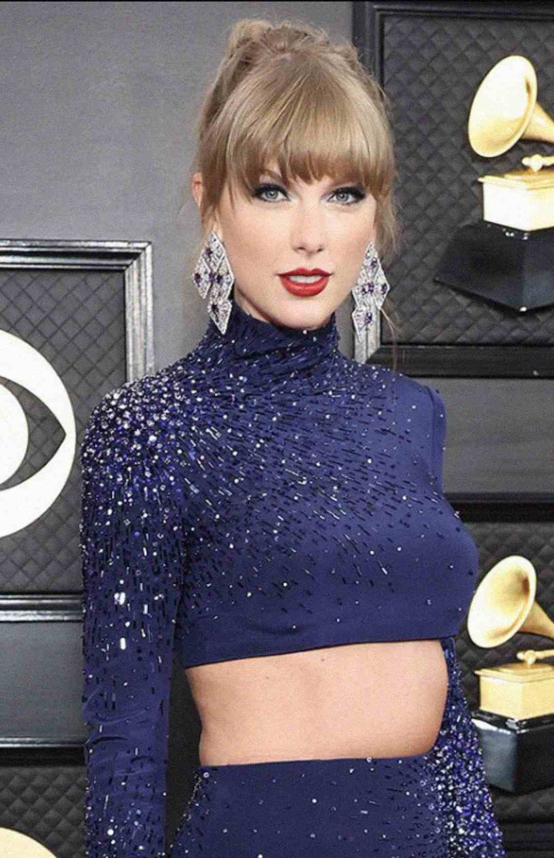 A woman with blonde hair in a high bun wears a sparkly, dark blue long-sleeve crop top and large diamond-shaped earrings, standing in front of Grammy Award trophies and a black backdrop.