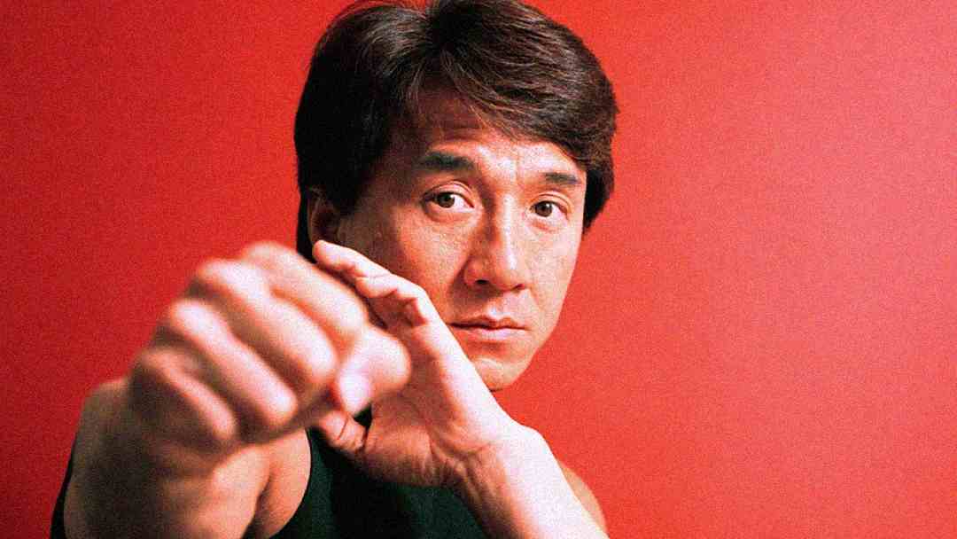 A man with short, dark hair poses in a martial arts stance against a plain red background, with one fist extended towards the camera and a serious expression on his face.