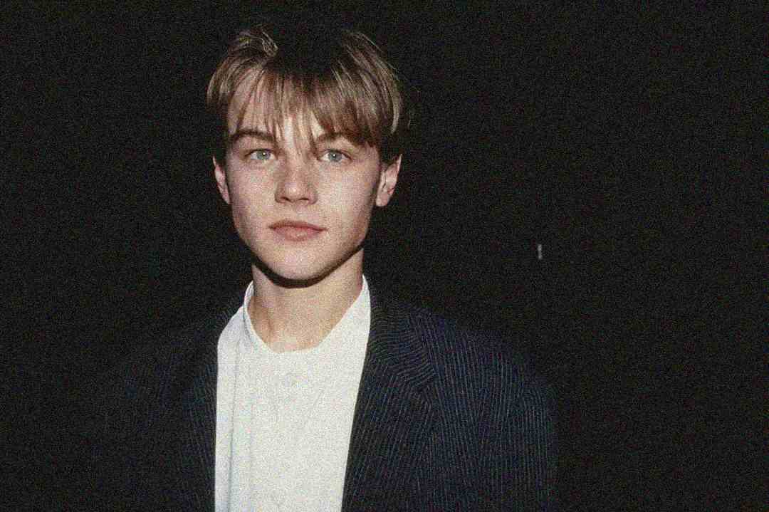 A young man with straight, light brown hair stands against a dark background, wearing a striped blazer over a white shirt, looking directly at the camera with a neutral expression.