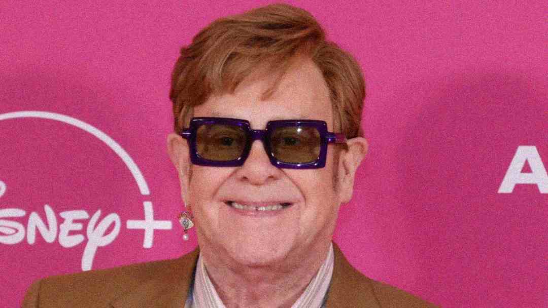 A smiling person with short light brown hair, wearing large square dark glasses, a brown jacket, and a white shirt stands in front of a pink Disney+ backdrop.