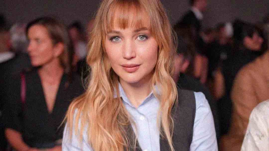 A woman with long blonde hair and bangs, wearing a light blue shirt and dark vest, sits indoors. The background is blurred with people seated behind her.