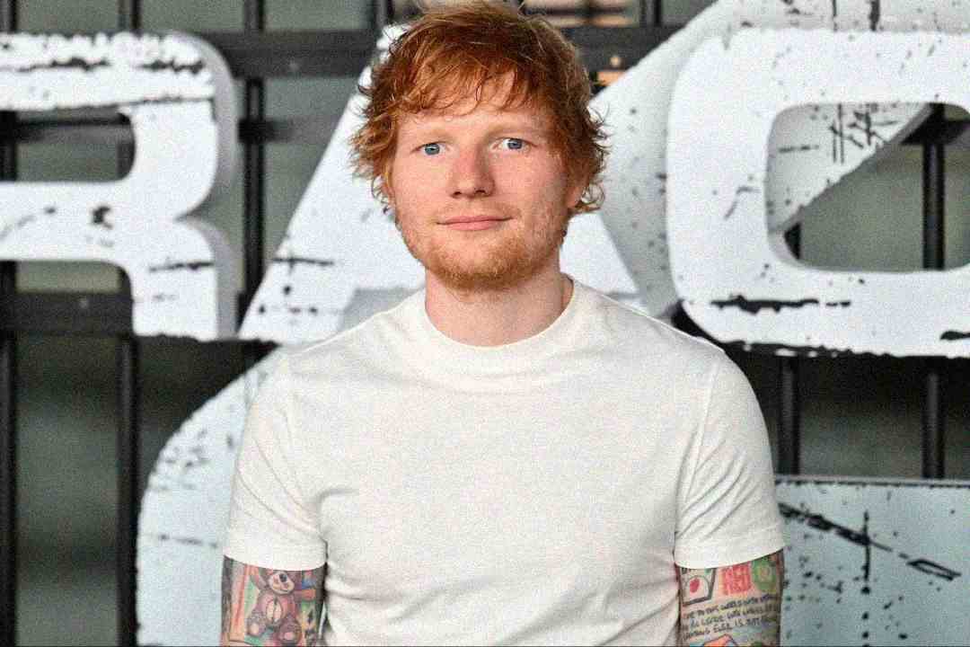 A man with red hair, a short beard, and tattoos on his arms stands in front of a large sign wearing a plain white t-shirt. The background features bold, partially visible letters.
