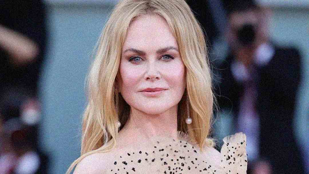 A woman with long blonde hair and light skin wears a beige, off-shoulder dress with black speckles. She has pearl drop earrings and a neutral expression, standing in front of a blurred background with people and cameras.