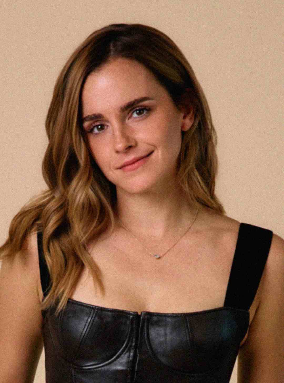 A woman with wavy light brown hair, wearing a black leather top and a delicate necklace, stands against a plain beige background, smiling softly at the camera.