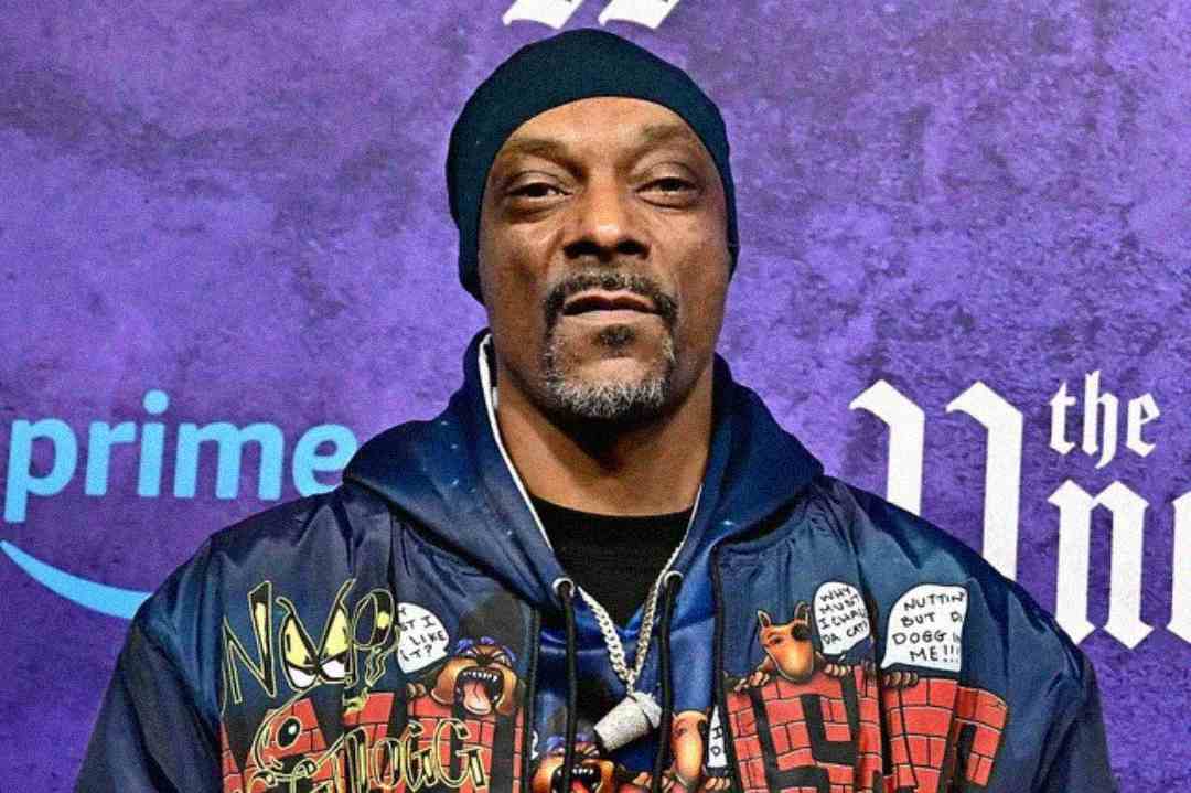 A man wearing a dark beanie and a jacket decorated with various pins and patches stands in front of a purple background with logos, including the Prime Video logo, visible behind him.