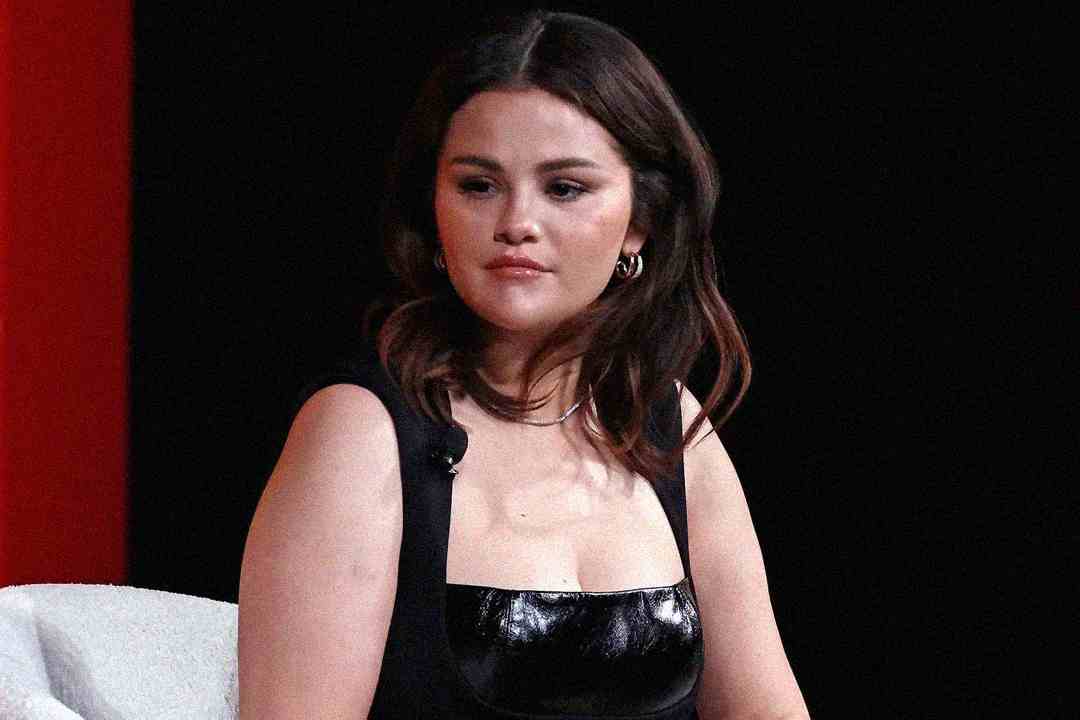 A woman with shoulder-length dark hair sits against a dark background, wearing a sleeveless black outfit with a shiny top and hoop earrings, looking slightly to the side with a neutral expression.
