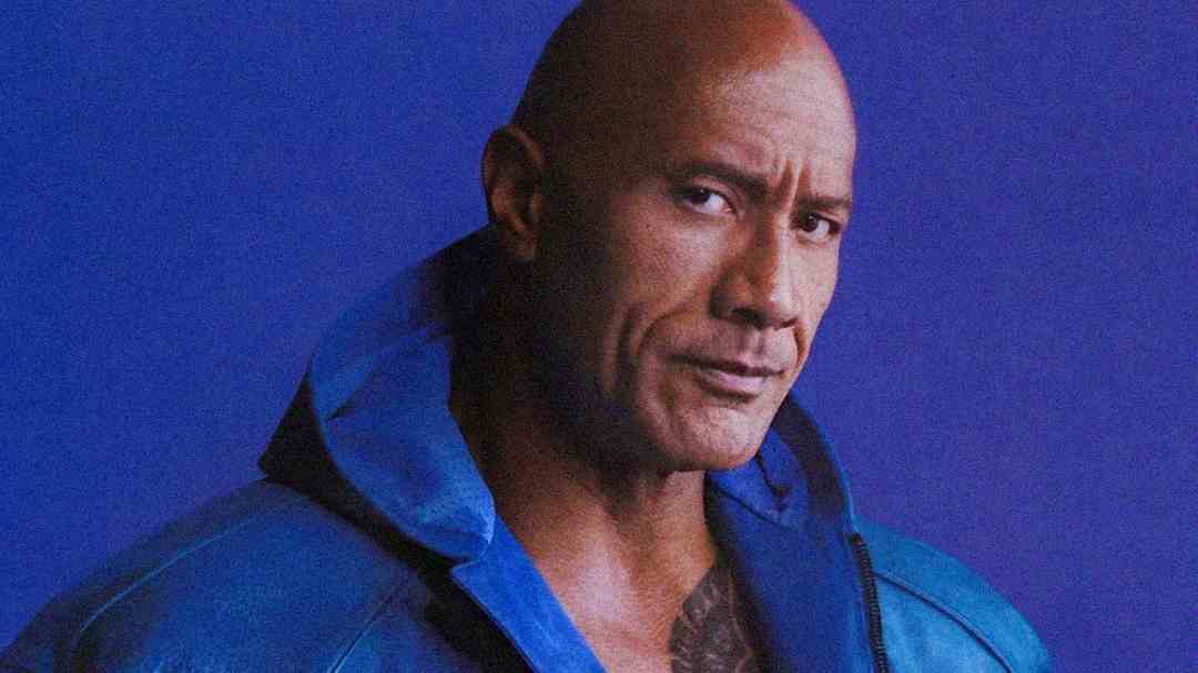A muscular, bald man wearing a blue jacket looks at the camera with a slight, serious expression, set against a plain purple background.