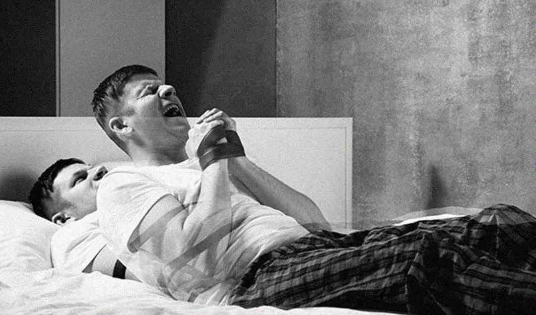 Two men sit on a bed; one leans back with his eyes closed, while the other, sitting in front, clasps his hands and appears to be singing or yelling, both wearing casual clothes in a monochrome setting.