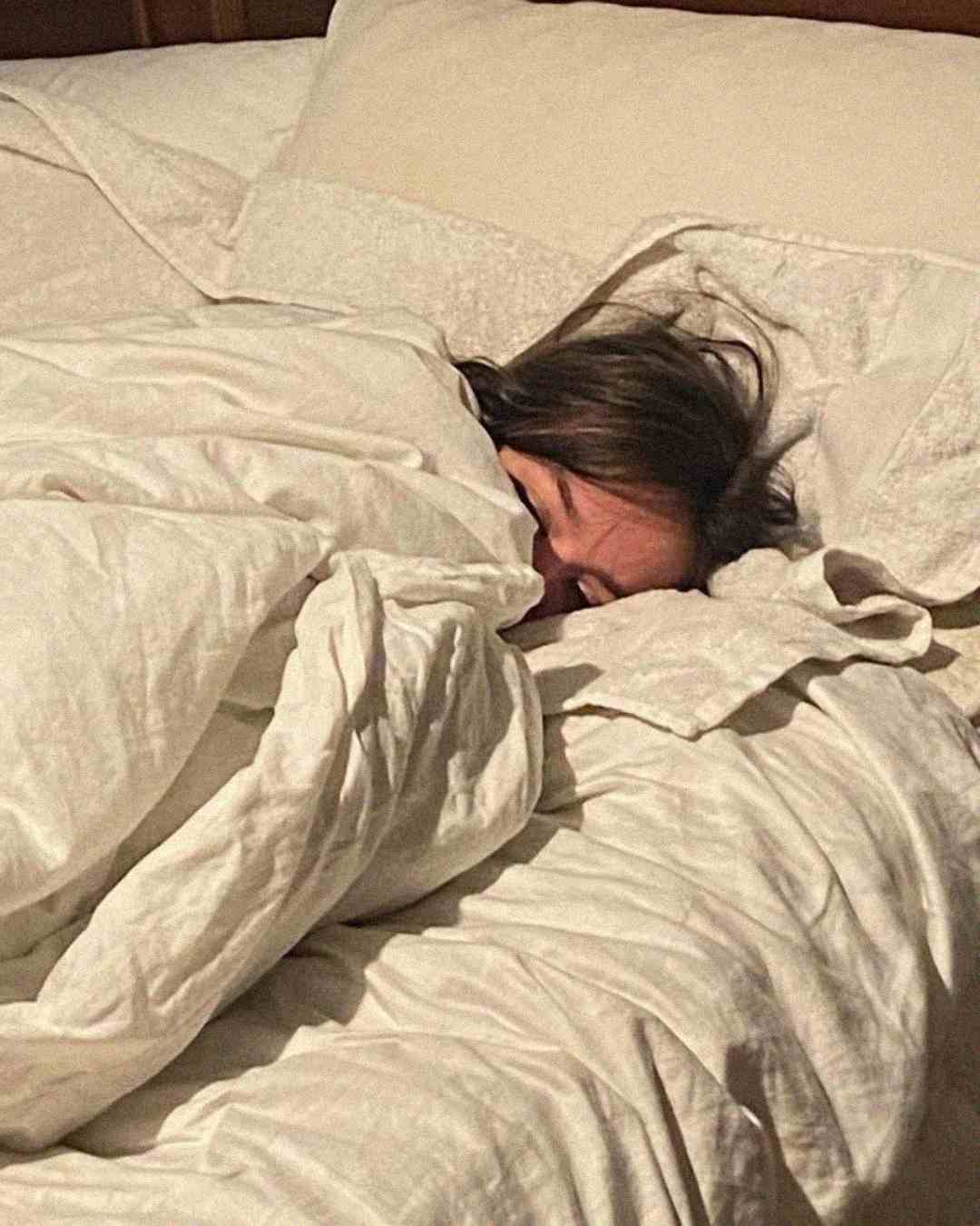 A person with brown hair is sleeping on a bed, mostly covered by a white comforter and pillow, with only part of their face visible.