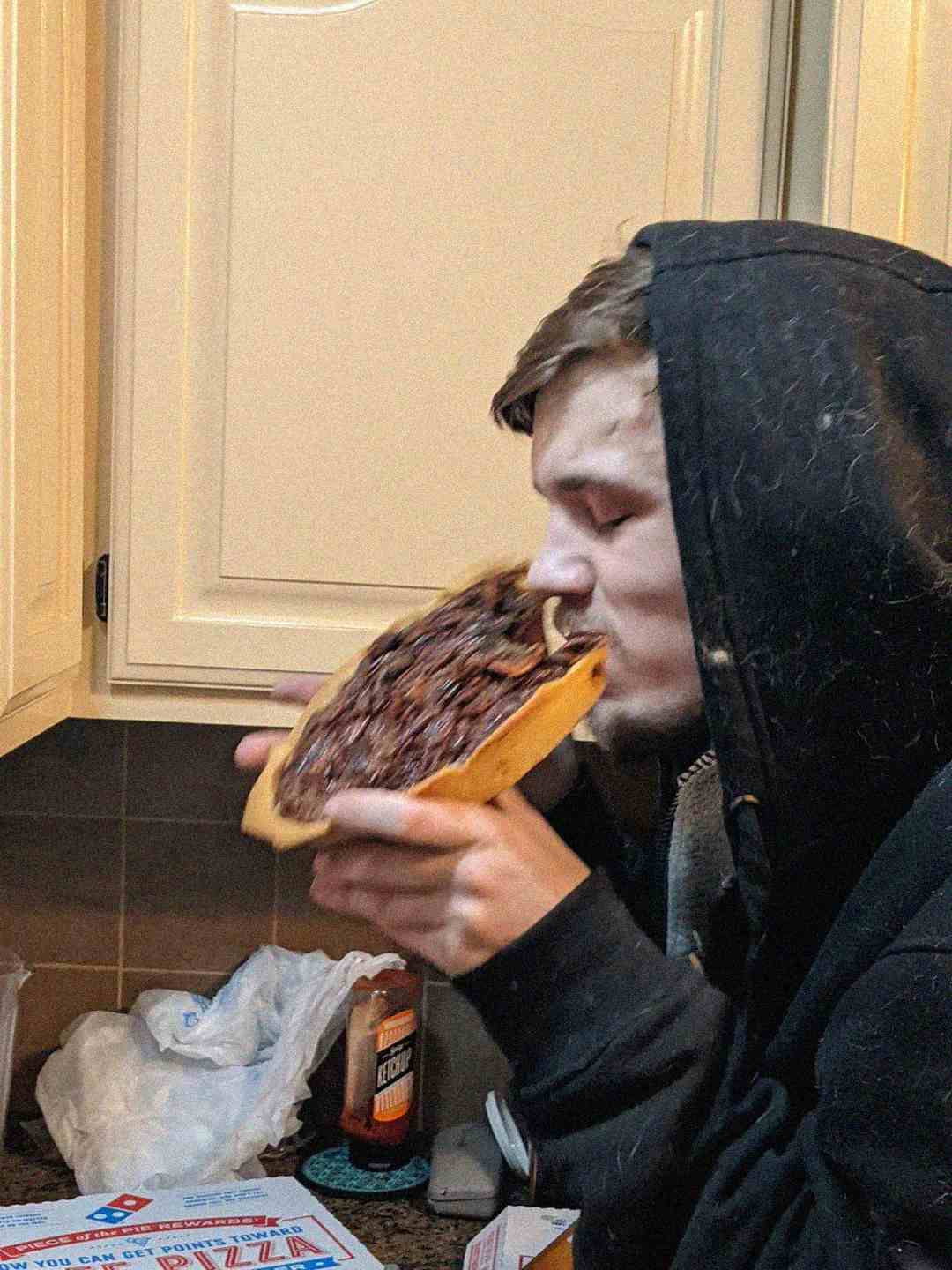 A person in a black hoodie is taking a big bite of a pizza slice covered with what appears to be chocolate or a dark spread, standing in a kitchen near a counter with various items.