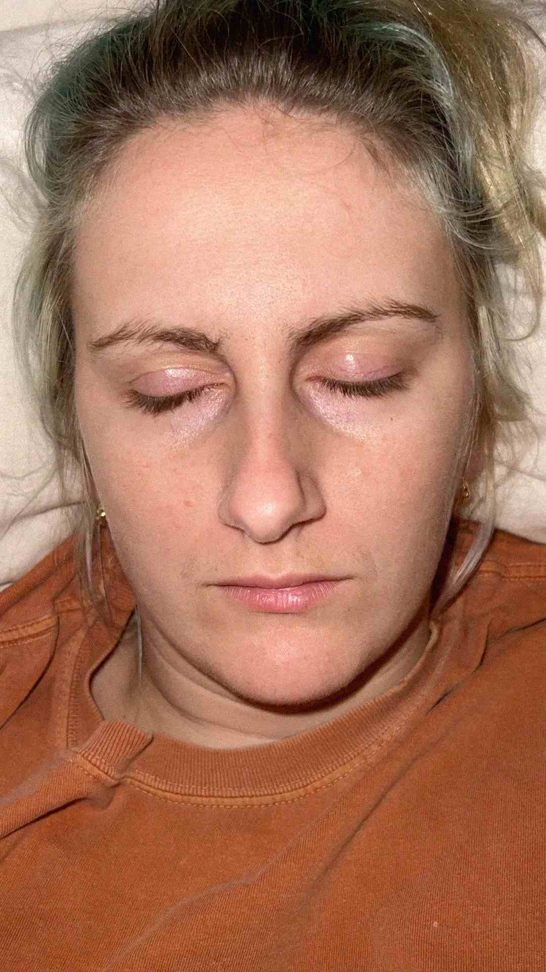 A person with closed eyes lying down, wearing an orange shirt. The lighting highlights their facial features and relaxed expression.