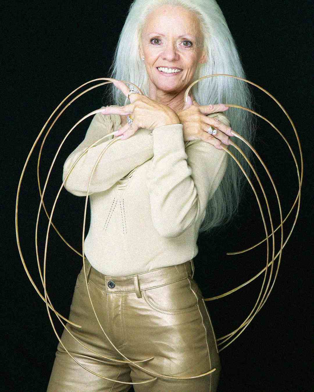 An older woman with long white hair smiles at the camera, crossing her arms over her chest and displaying extremely long, curved fingernails. She wears a beige top and metallic pants, with a black background.