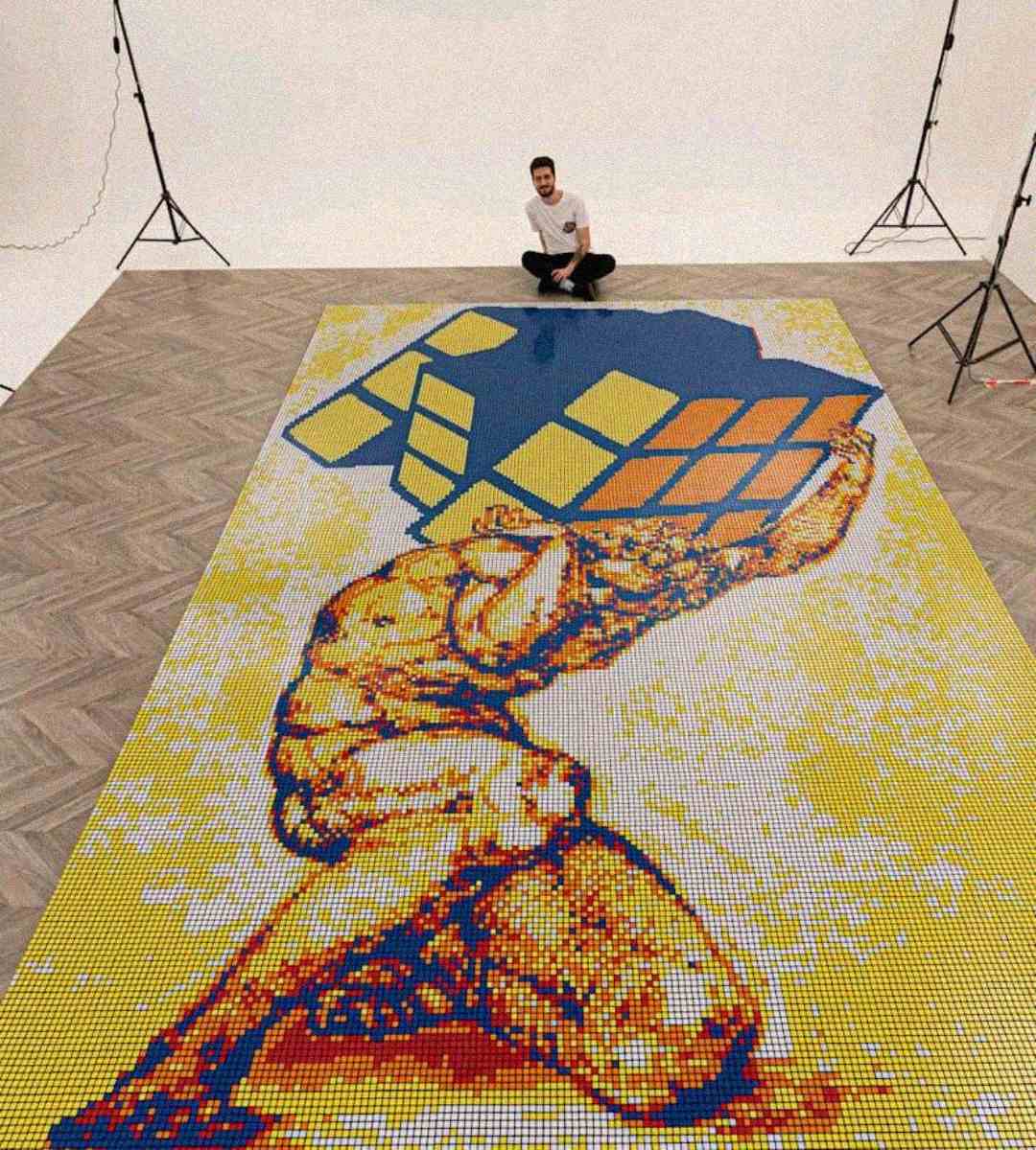A man kneels behind a large floor mosaic of Atlas holding a Rubik's Cube instead of the world, made from many small colored tiles, with studio lights in the background.