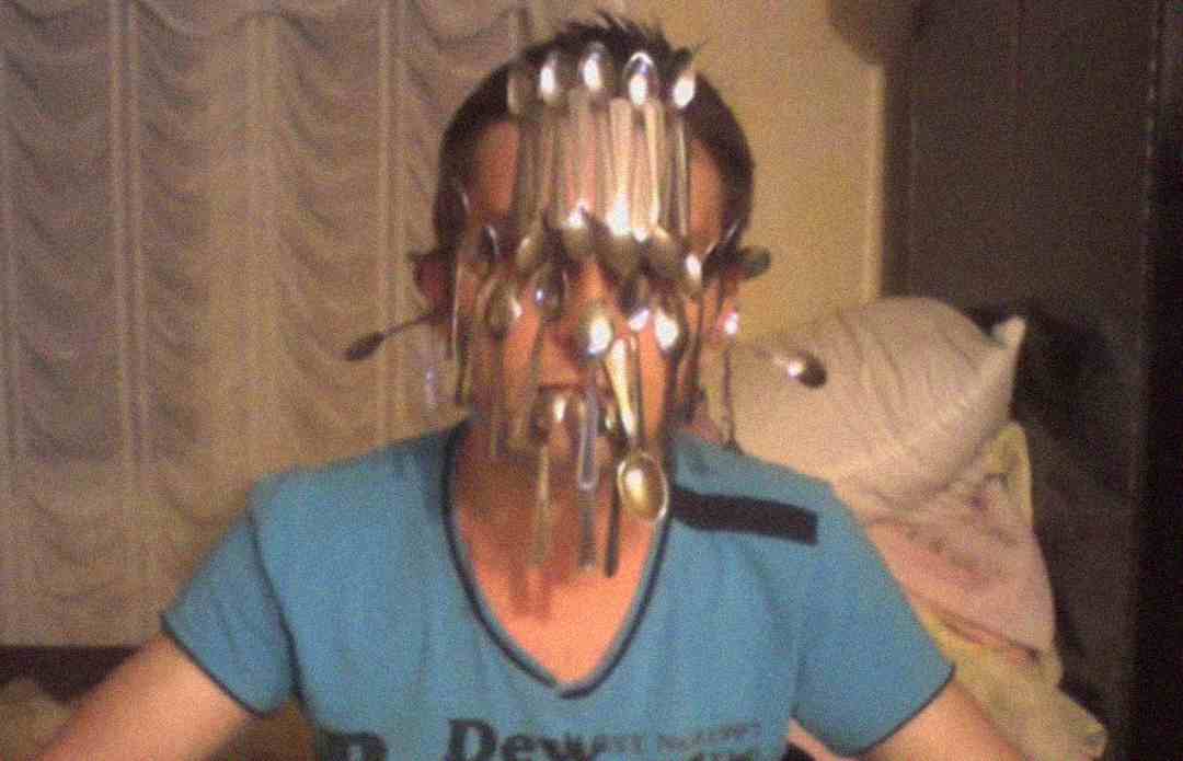 A person wearing a blue shirt sits indoors with numerous metal spoons hanging from and sticking to their face, obscuring their features. The background shows curtains and a pile of blankets on a couch.