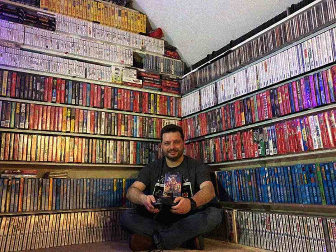 A man sits cross-legged on the floor in a room filled with shelves of video game cases and boxes, smiling and holding a game controller and a game case.