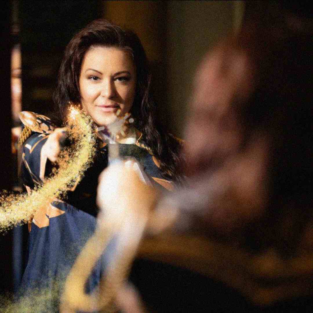 A woman in a dark robe uses a wand to cast golden sparkles, conjuring magic in front of her. She looks confident and focused, and the magical effect glows brightly in the scene.