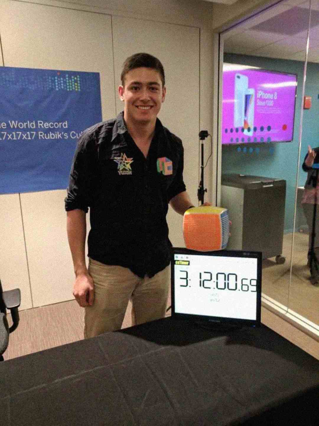 A smiling man stands next to a digital timer displaying "3:12:00.69," holding a large Rubik's Cube. A banner behind him mentions a world record for solving a 17x17x17 Rubik’s Cube.