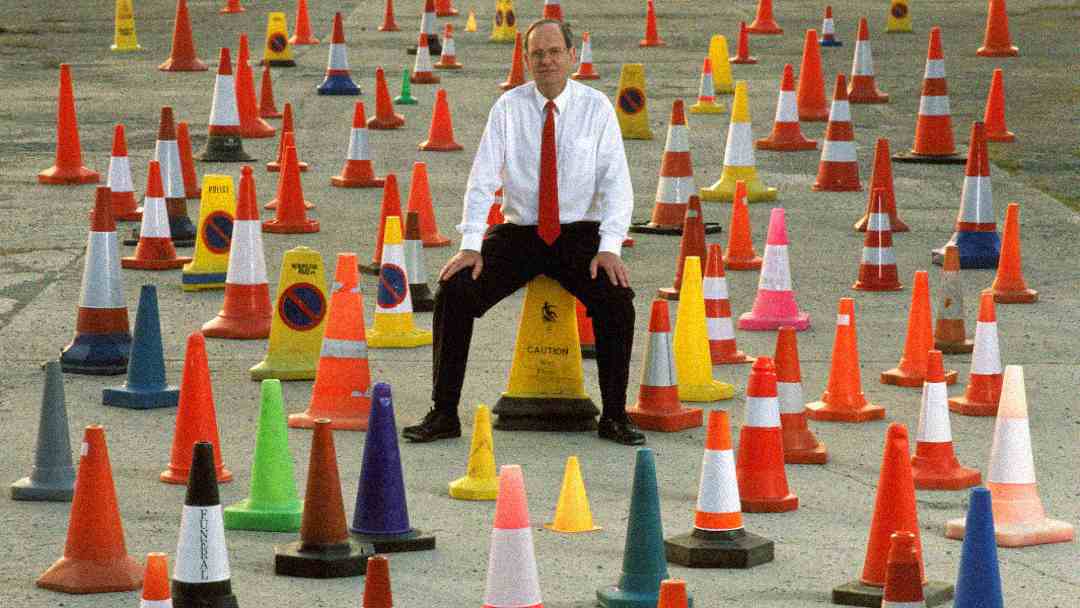A man in a shirt and tie sits on a yellow caution cone surrounded by dozens of colorful traffic cones and warning signs on an outdoor pavement.