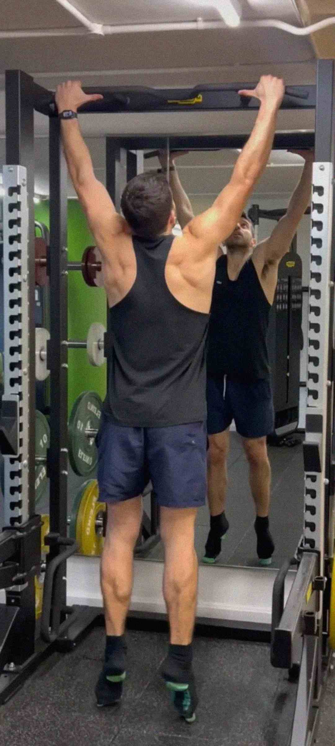 A man wearing a black tank top and navy shorts performs a pull-up in a gym, standing in front of a mirror and power rack with weight plates visible on the side.