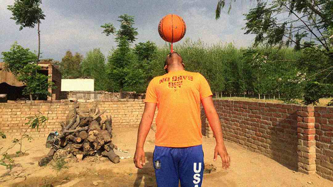 A person in an orange shirt and blue pants balances a basketball on their face, standing in a sandy yard with trees, a brick wall, and a woodpile in the background.
