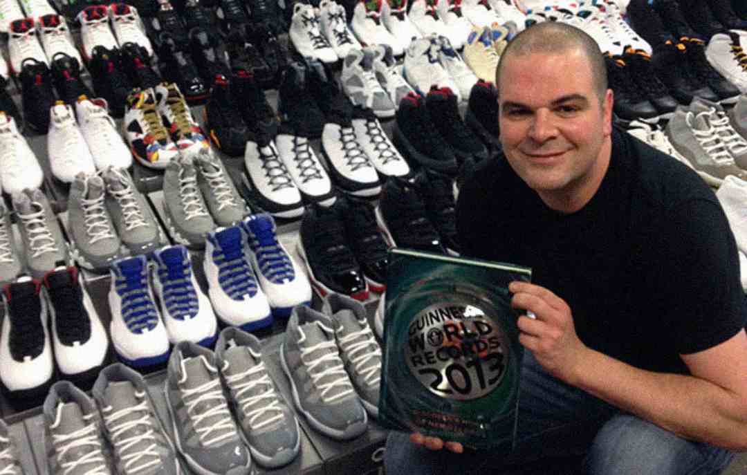 A man smiles while holding a Guinness World Records 2013 plaque, sitting in front of an extensive collection of sneakers arranged in rows.
