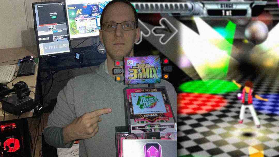 A person wearing glasses stands indoors, pointing at a stack of Dance Dance Revolution games with arcade graphics in the background and computer monitors nearby.