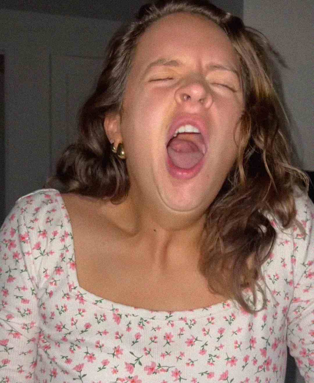 A woman with wavy brown hair, wearing a white floral top, is yawning with her mouth wide open and eyes closed. She appears to be tired or sleepy in a softly lit indoor setting.