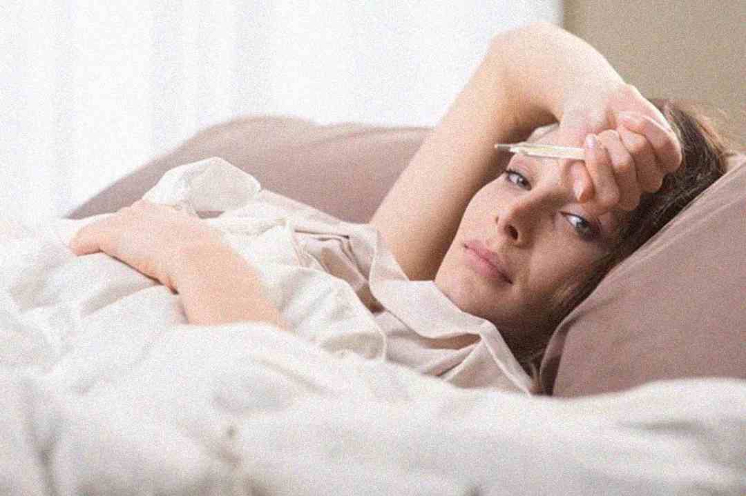 A woman lies in bed looking unwell, holding a thermometer to her forehead, partially covered by a white blanket, in a softly lit room.