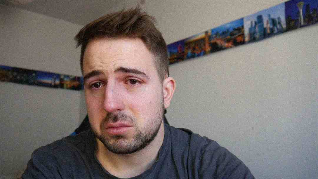 A young man with short brown hair and facial hair sits indoors, looking sad and teary-eyed, with a somber expression. There are colorful photos arranged in a strip on the wall behind him.