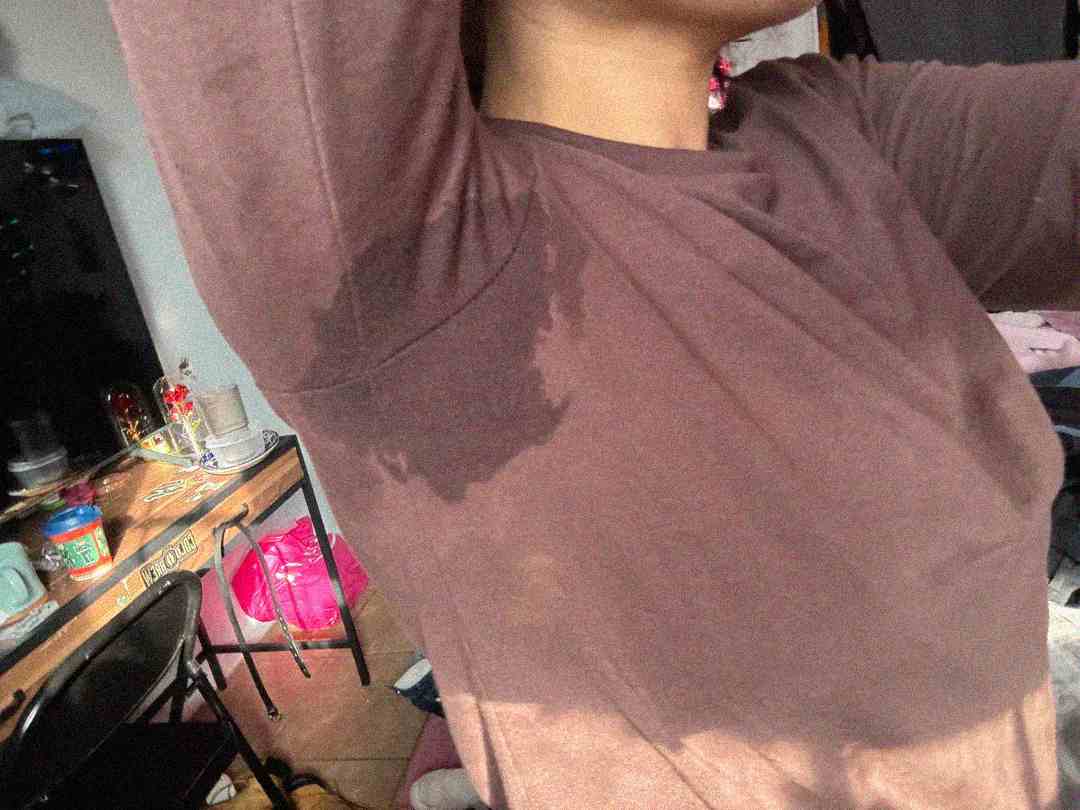 A person wearing a brown long-sleeve shirt lifts their arm, revealing a large sweat stain under the armpit. The background shows a cluttered desk, a chair, and a pink bag on the floor.