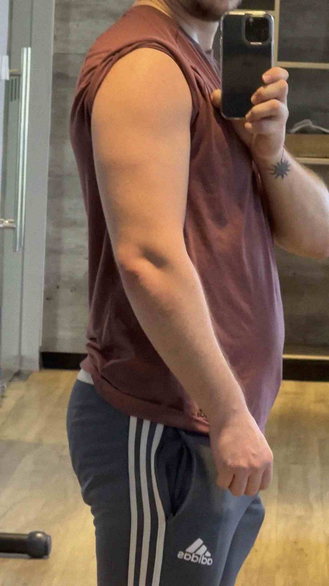 A person wearing a sleeveless maroon shirt and gray Adidas pants takes a mirror selfie in a gym, showing their flexed arm and side profile.