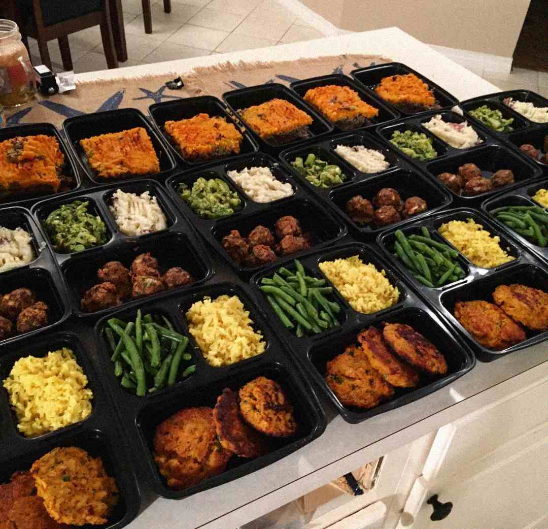 A kitchen counter covered with multiple black meal prep containers, each filled with portions of cooked food such as green beans, yellow rice, meatballs, pasta, and patties, arranged in neat rows.